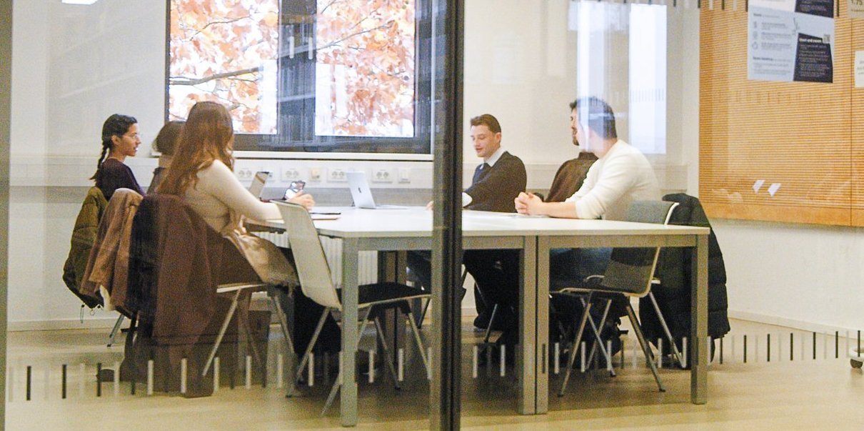 Vier Studierende sitzen an einem Tisch in einem modernen, hellen Besprechungsraum mit Glaswänden und diskutieren.