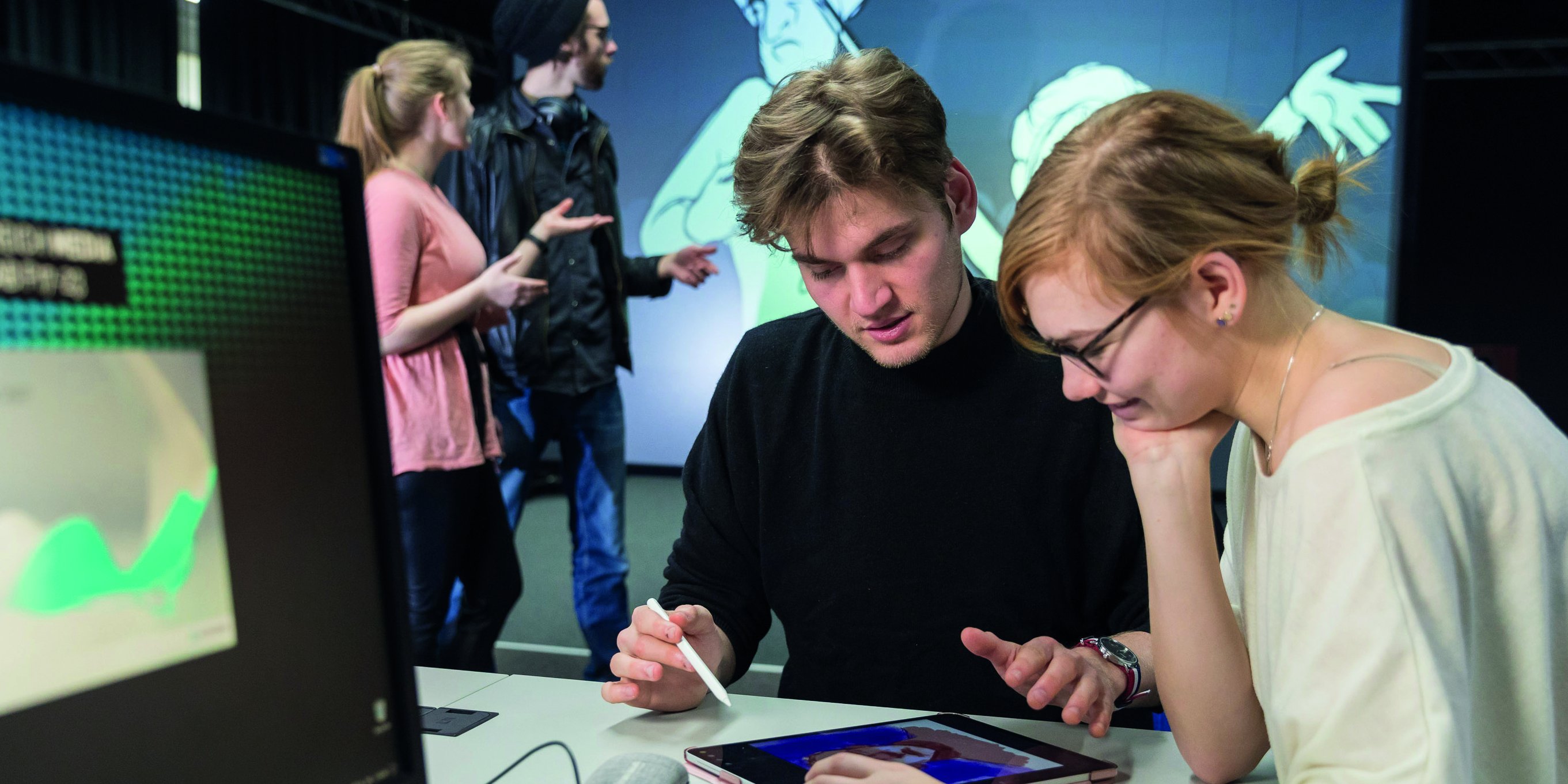 zwei Studierende sitzen an einem Tisch und blicken gemeinsam auf ein Tablet, im Hintergrund stehen zwei Studierende vor einem BIldschirm