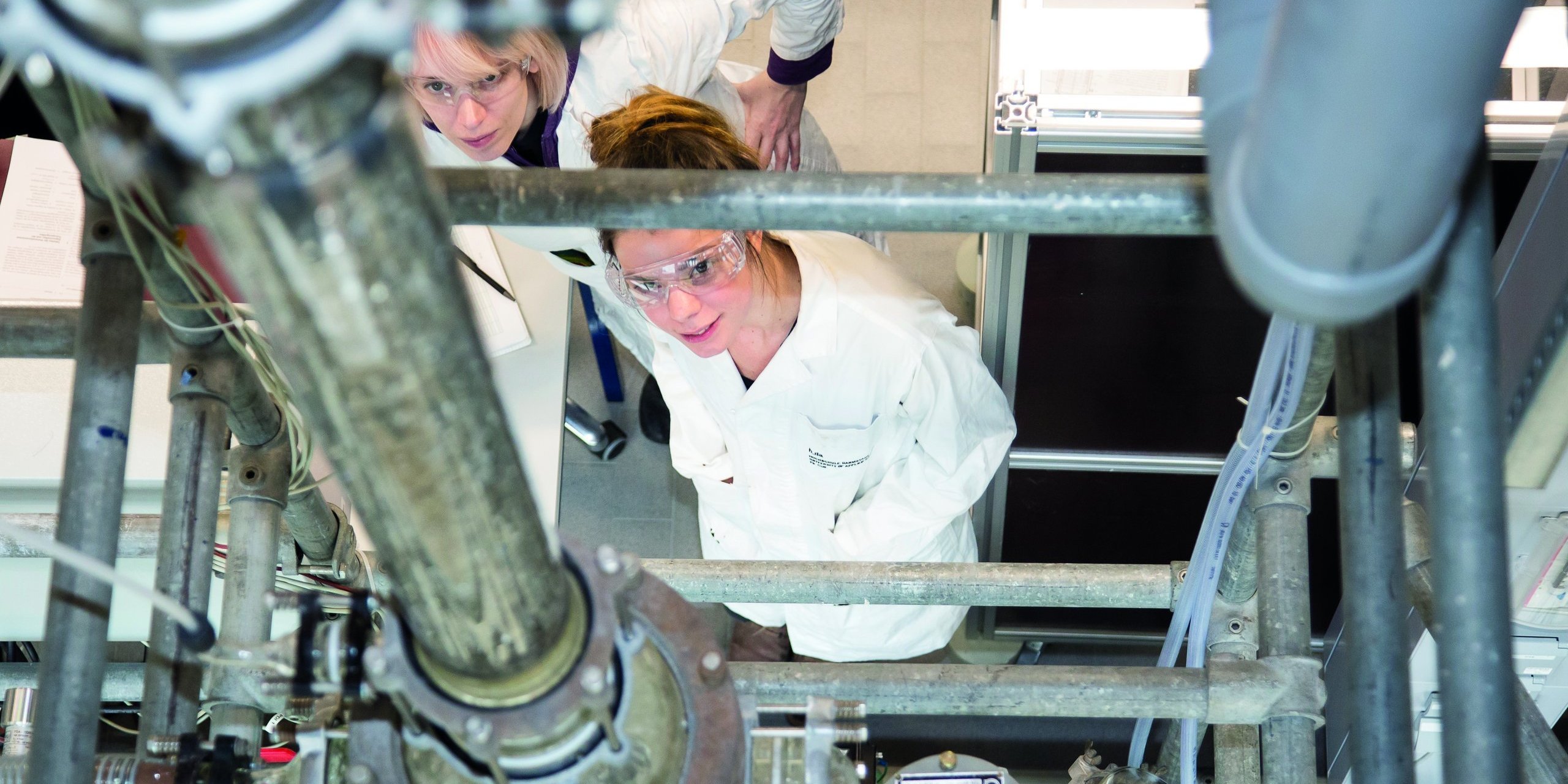 Two people in lab coats with safety goggles look into a plant from below.