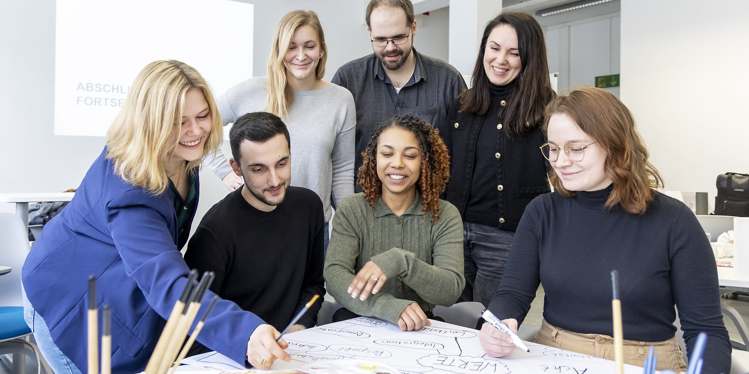 mehrere Personen mit Stiften an einem Tisch, auf dem ein großer Papierbogen liegt 