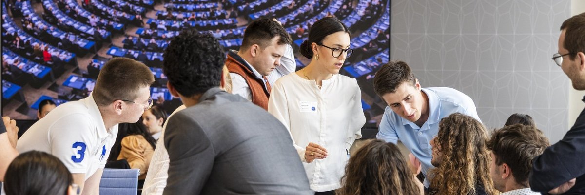 Gruppe von Studierenden diskutiert vor einem großen Screen, auf dem der Plenarsaal des EU-Parlaments zu sehen ist. 