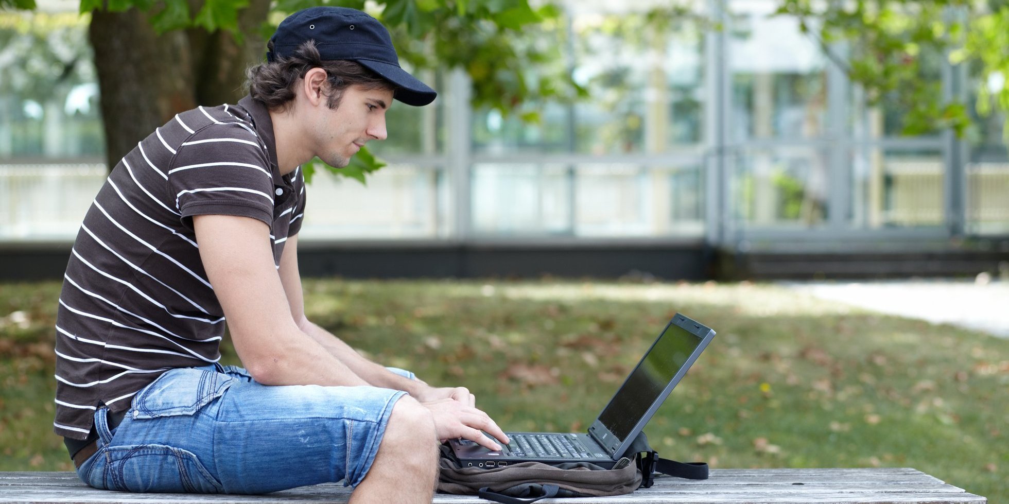 Ein Student sitzt auf einer Bank im Freien auf dem Campus und tippt auf seinem Laptop.