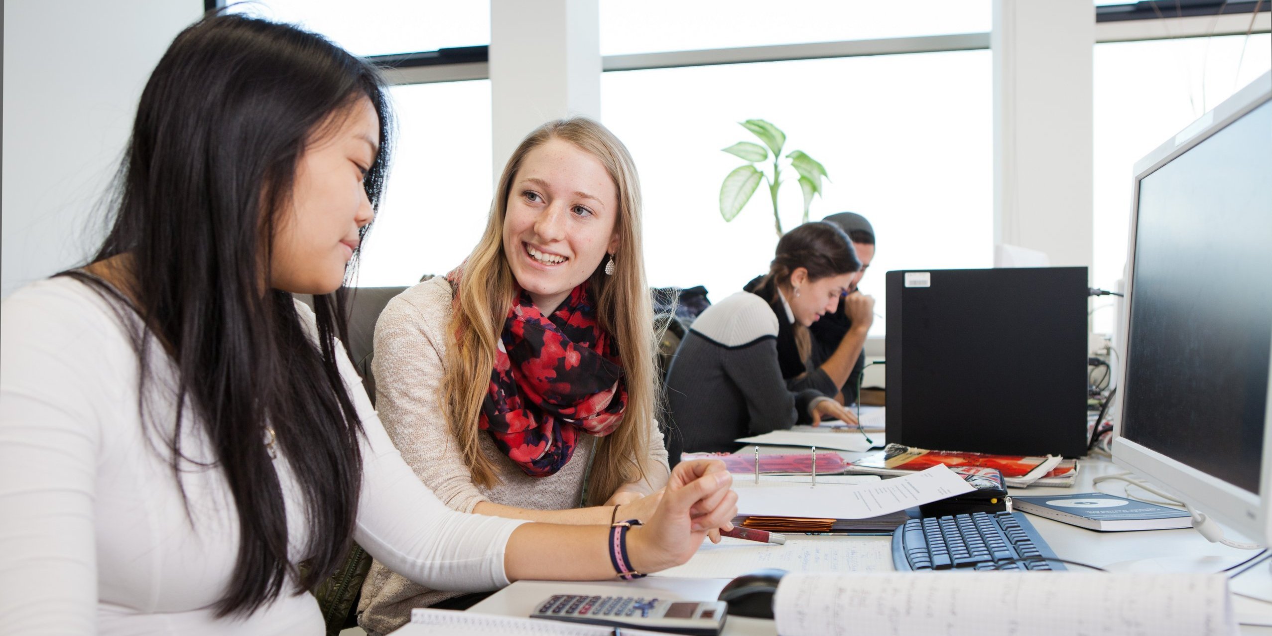 Two female students sTwo people sitting at a table with calculator, computer and writing utensilsit at a table with a calculator, computer and writing utensils.