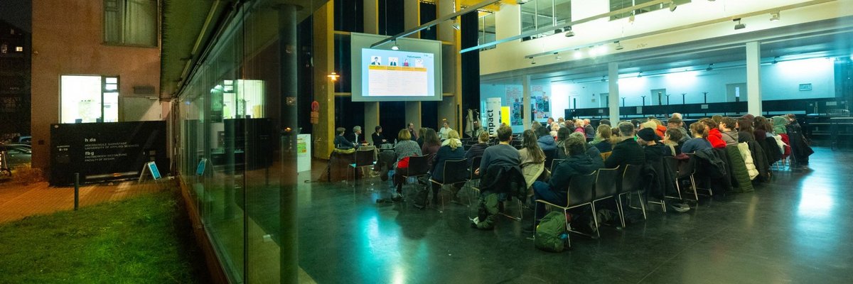 Blick durch eine Glasfront in den erleuchteten Veranstaltungsraum am Fachbereich Gestaltung mit Stuhlreihen und Publikum