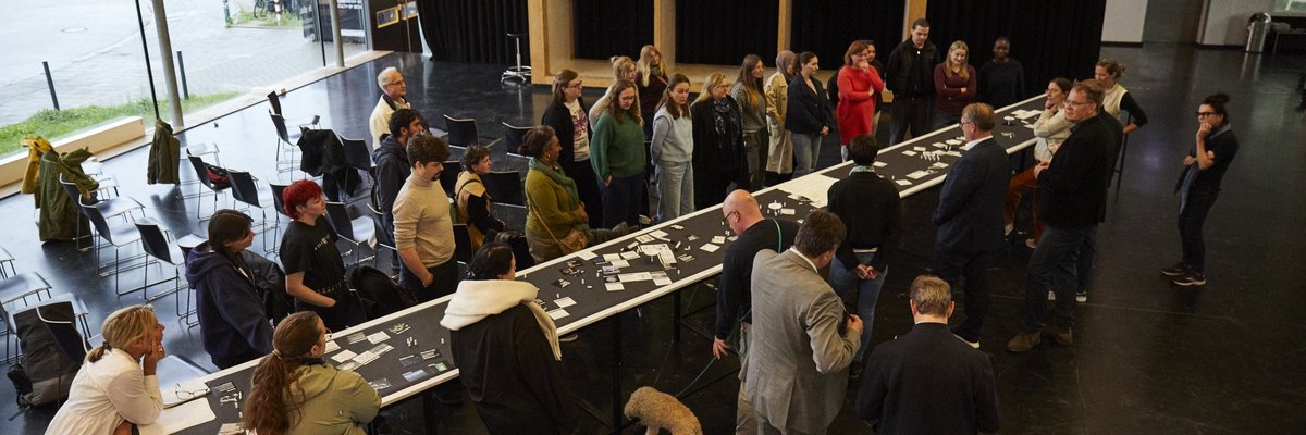 Workshopteilnehmende stehen beidseitig an einem langen Tisch im Foyer des Fachbereichsgebäudes Gestaltung auf der Mathildenhöhe