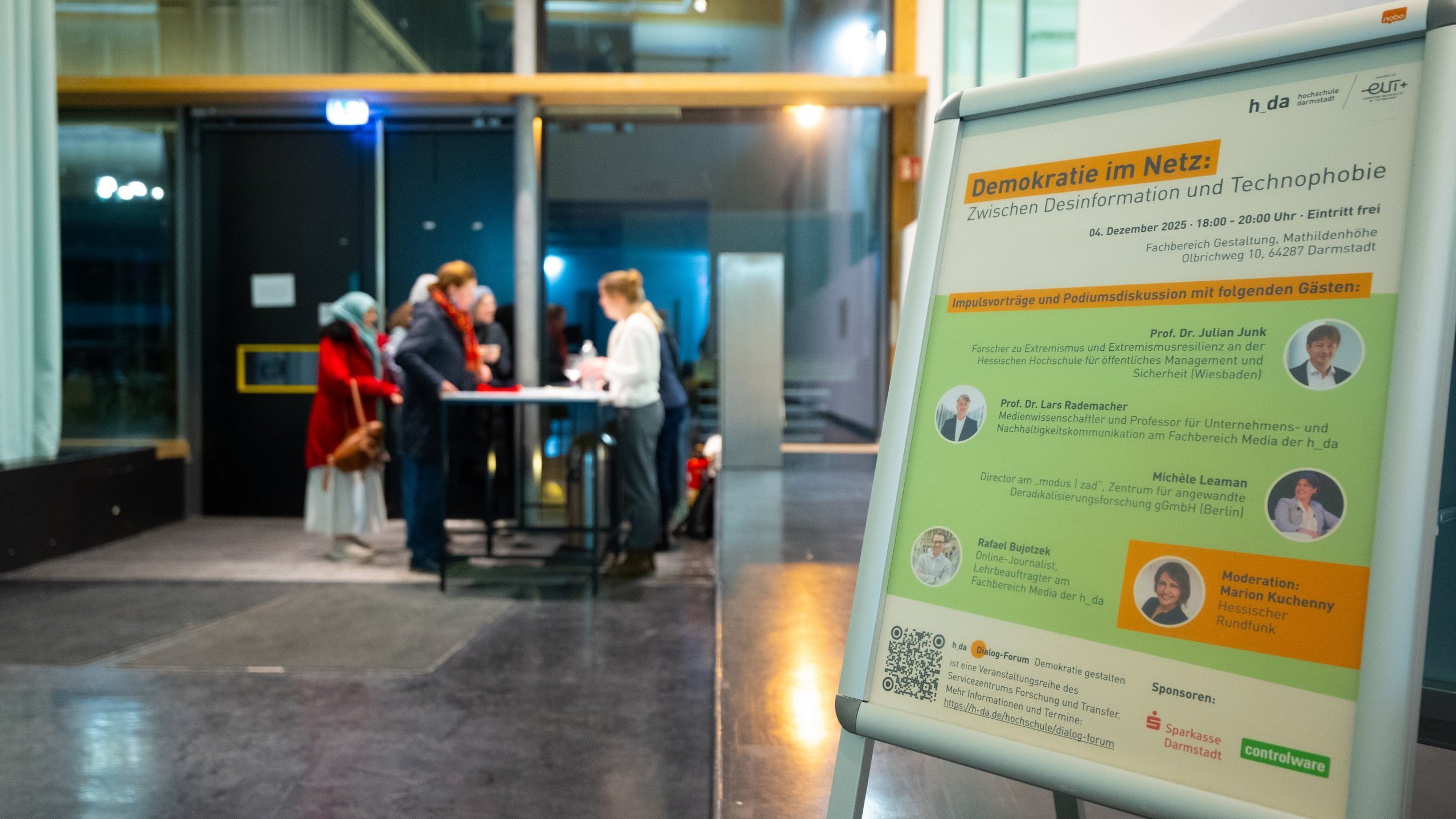 Gäste kommen im Gebäude des Fachbereichs Gestaltung an; im Vordergrund ein Plakat des 6. h_da Dialog-Forums