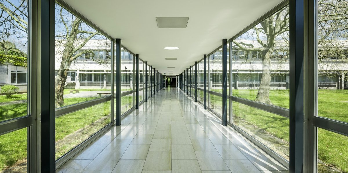 Campus Dieburg, ein langer gläserner Verbindungsgang, der zu einem entfernt liegenden Gebäude führt. Links und rechts außen Wiesen mit Bäumen und Sitzplätzen