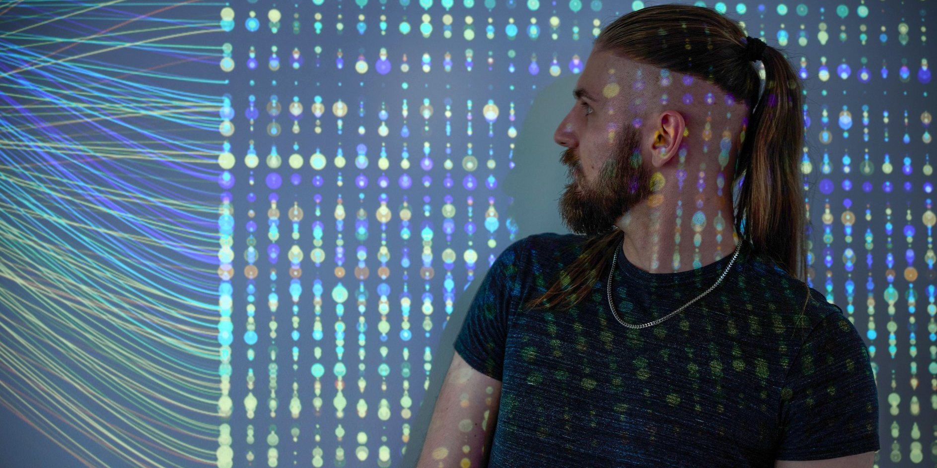 Mann mit Zopf und Bart steht seitlich vor einer Wand mit digital wirkenden, farbigen Datenpunkten und Linien.