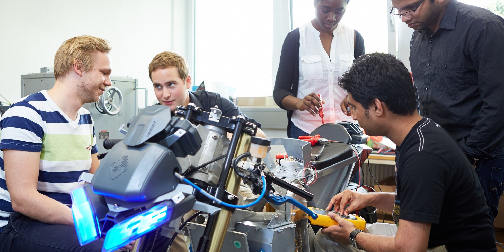 five people, equipped with measuring instruments and tools, are gathered around an electric motorbike