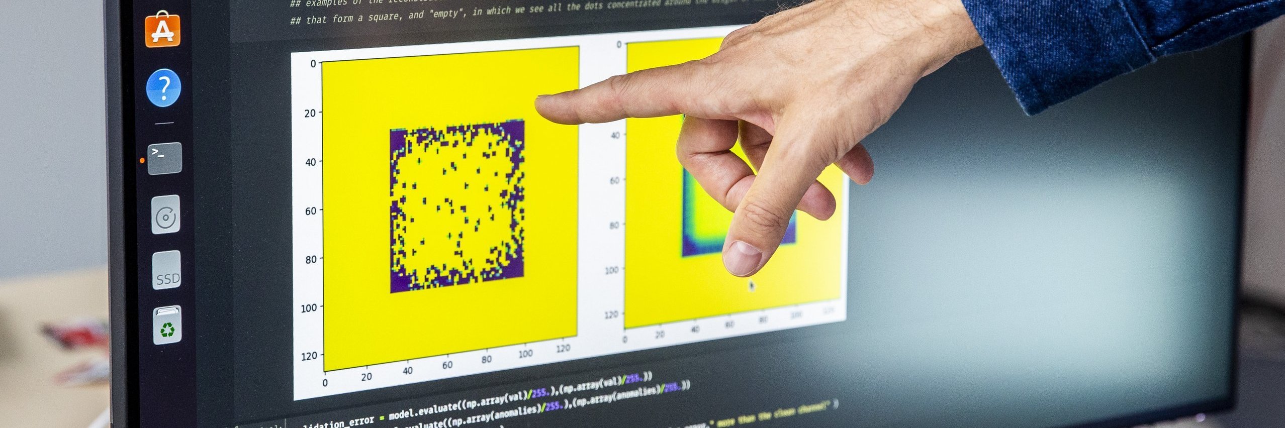 Ein Finger deutet auf einen Monitor. Auf dem Monitor ist ein Quellcode und eine gelbe Grafik zu sehen.