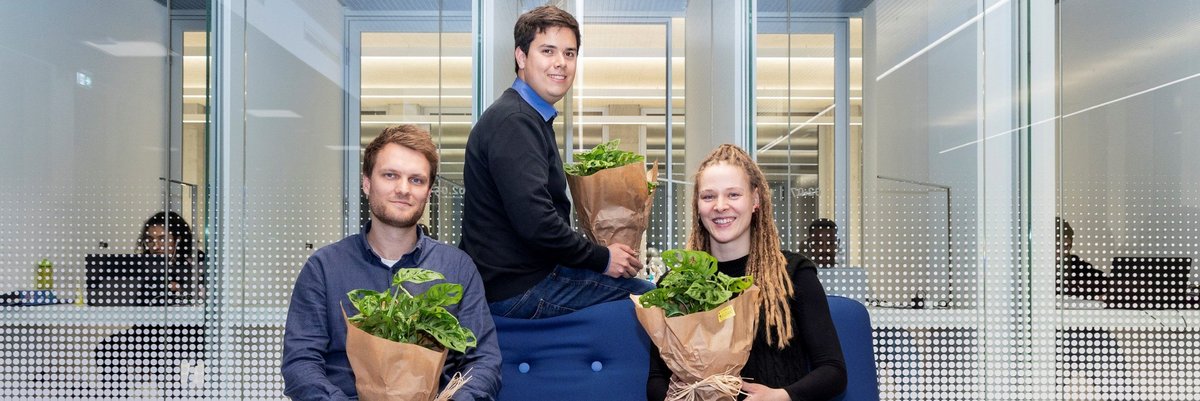 Das Foto zeigt drei der mit Preisen ausgezeichneten Studierenden sitzend auf einem Sofa und der Sofa-Rückenlehne. Alle drei halten eine Grünpflanze als Geschenk in den Händen.