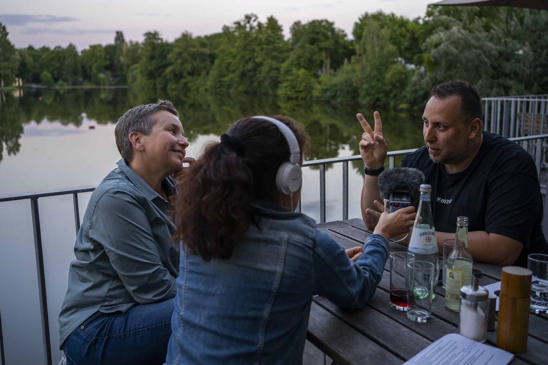 Nicole Saenger, DJ Ufuk und Christina Janssen sitzen auf der Terrasse des Cafes am Woog und diskutieren. Im Hintergrund der See. Abendstimmung. 