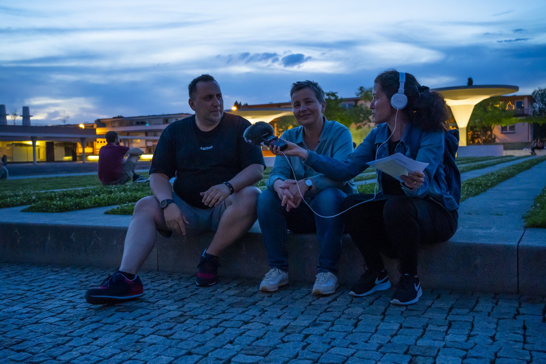 DJ Ufuk, Christina Janssen und Nicole Saenger sitzen nebeneinander auf einem Mäuerchen auf dem nächtlichen Büchnerplatz. 